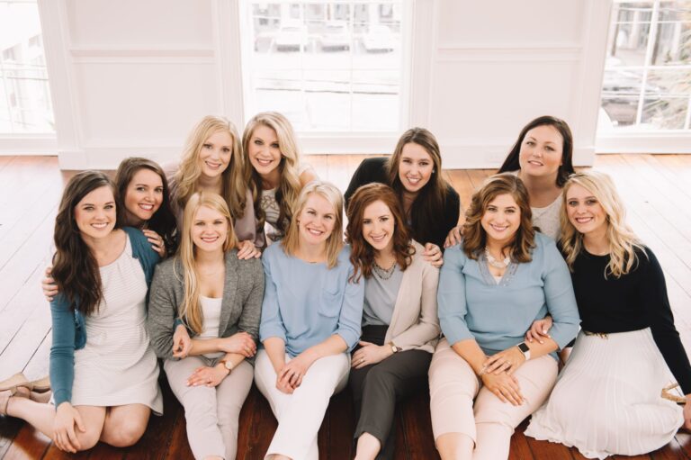 A group of women sit together on a wooden floor, smiling