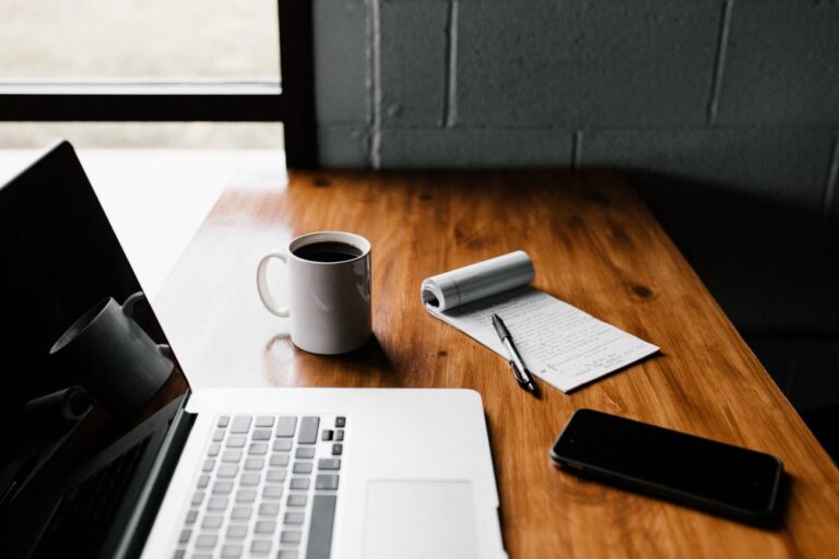 laptop on a wood desk with a notebook and coffee mug