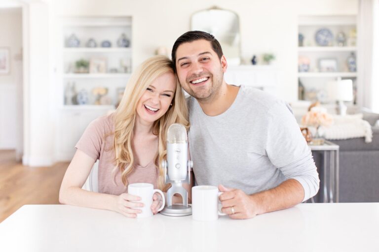 A man and woman smile with their heads together, seated behind a kitchen island. A podcasting microphone sits between them on the counter.