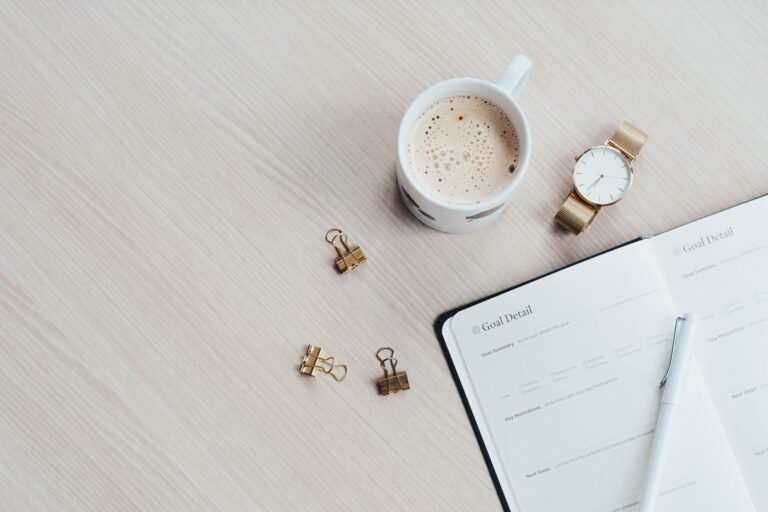 A goal planner sits on a table surrounded by a cup of coffee, watch, and binder clips