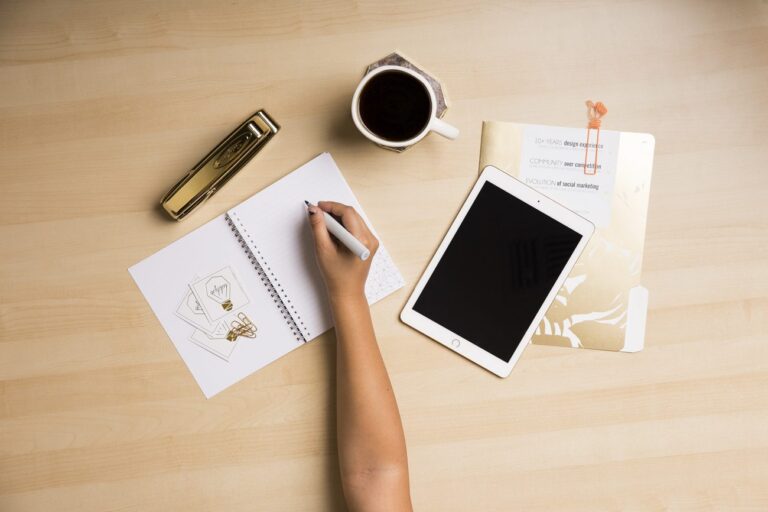 a desk with a person writing next to a tablet and cup of coffee