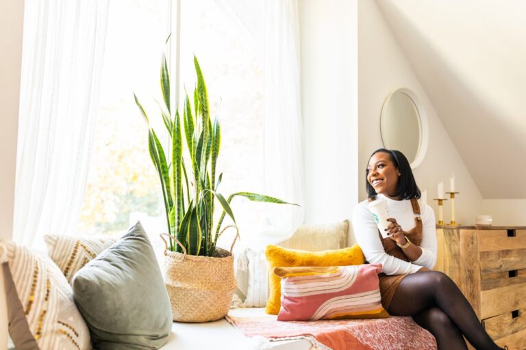 woman sitting on a couch by a large window with plants and blankets