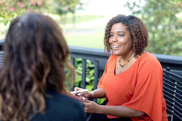 A woman in an orange shirt sits at a table outside, conversing with another person facing away from camera